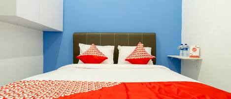 A bed with a white and red bedspread, two red pillows, and a headboard. There is a shelf with bottled water and a box of tissues.