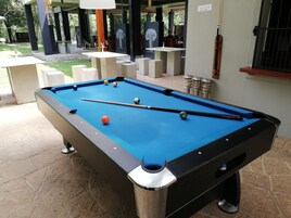 Une table de billard avec des billes et des queues, entourée de tables et de chaises, dans un espace extérieur où un panneau indique que l’accès est interdit aux personnes de moins de 22 ans.