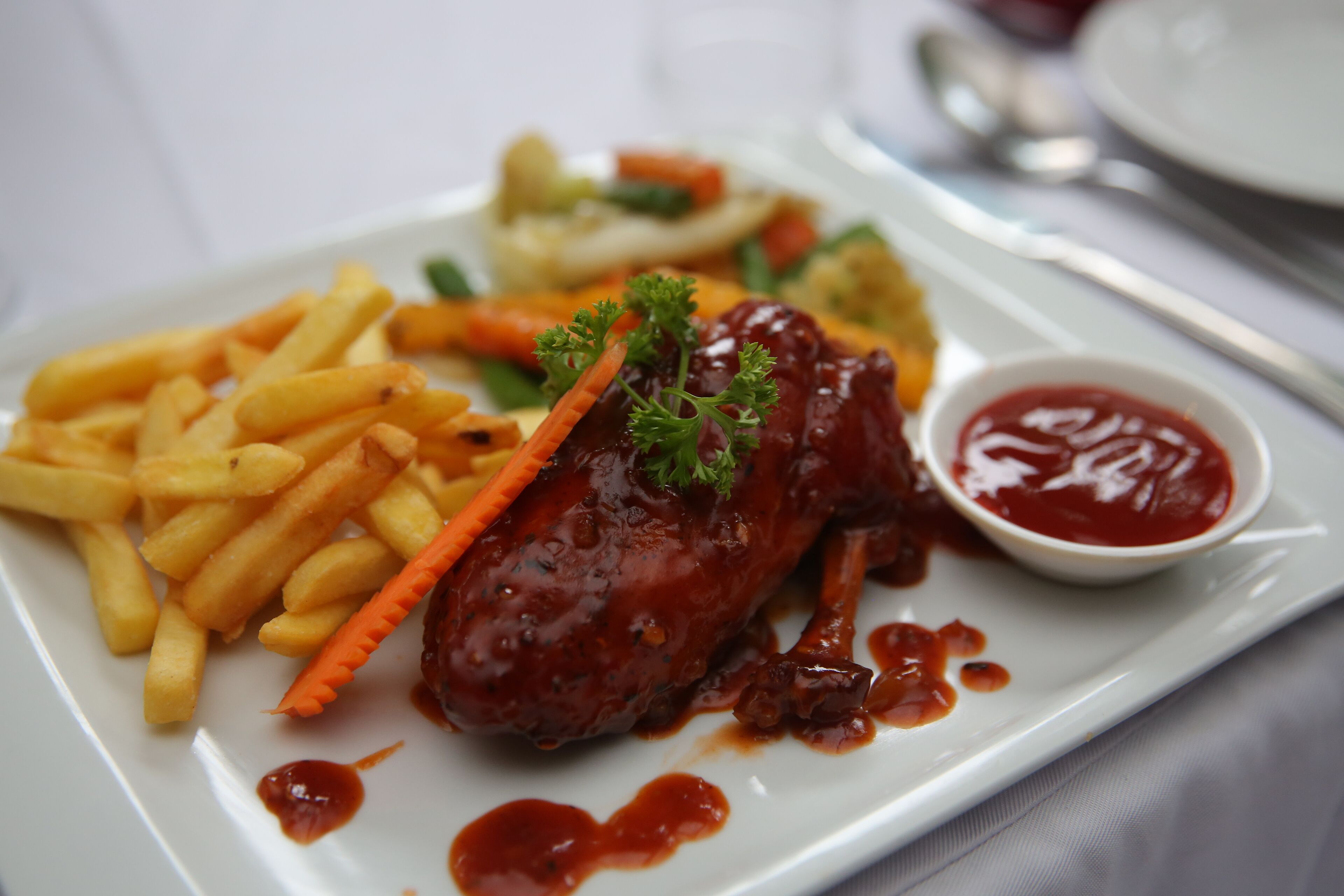 A plate with a glazed meat dish, French fries, a carrot garnish, and a small bowl of ketchup.