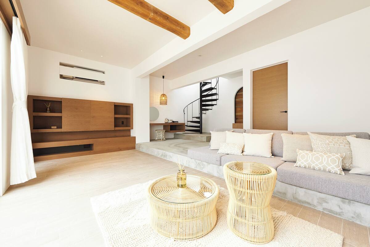 Een moderne woonkamer met een trap, houten balken, een bank en twee ronde bijzettafels van riet.
