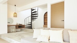 Uma sala de estar moderna com um sofá de cor clara, um espelho redondo, uma mesa lateral de vime e uma escada espiral preta que leva a um andar superior.