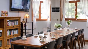 A dining area with a long table set for tea, surrounded by chairs, a television mounted on the wall, and a bookshelf with books. - The Hue Largo Bundang (Yongin)