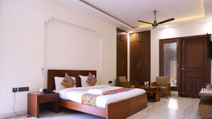 A hotel room with a large bed, a bedside table, a chair, a desk, a wardrobe, and a ceiling fan. - Kumar Residency (Noida)
