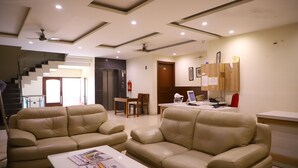 A modern living room with two beige sofas, a coffee table with newspapers, a dining area with a table and chairs, and a kitchen area with wooden cabinets and a refrigerator. - Kumar Residency (Noida)