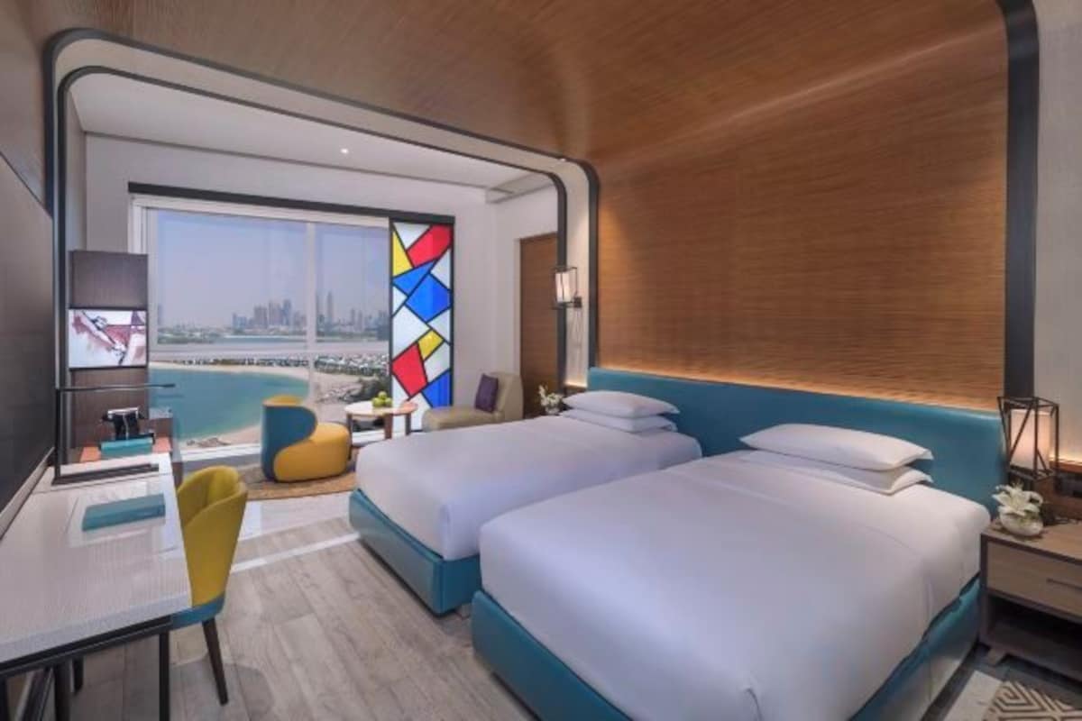 A modern hotel room with two beds, a desk, and a view of the city.