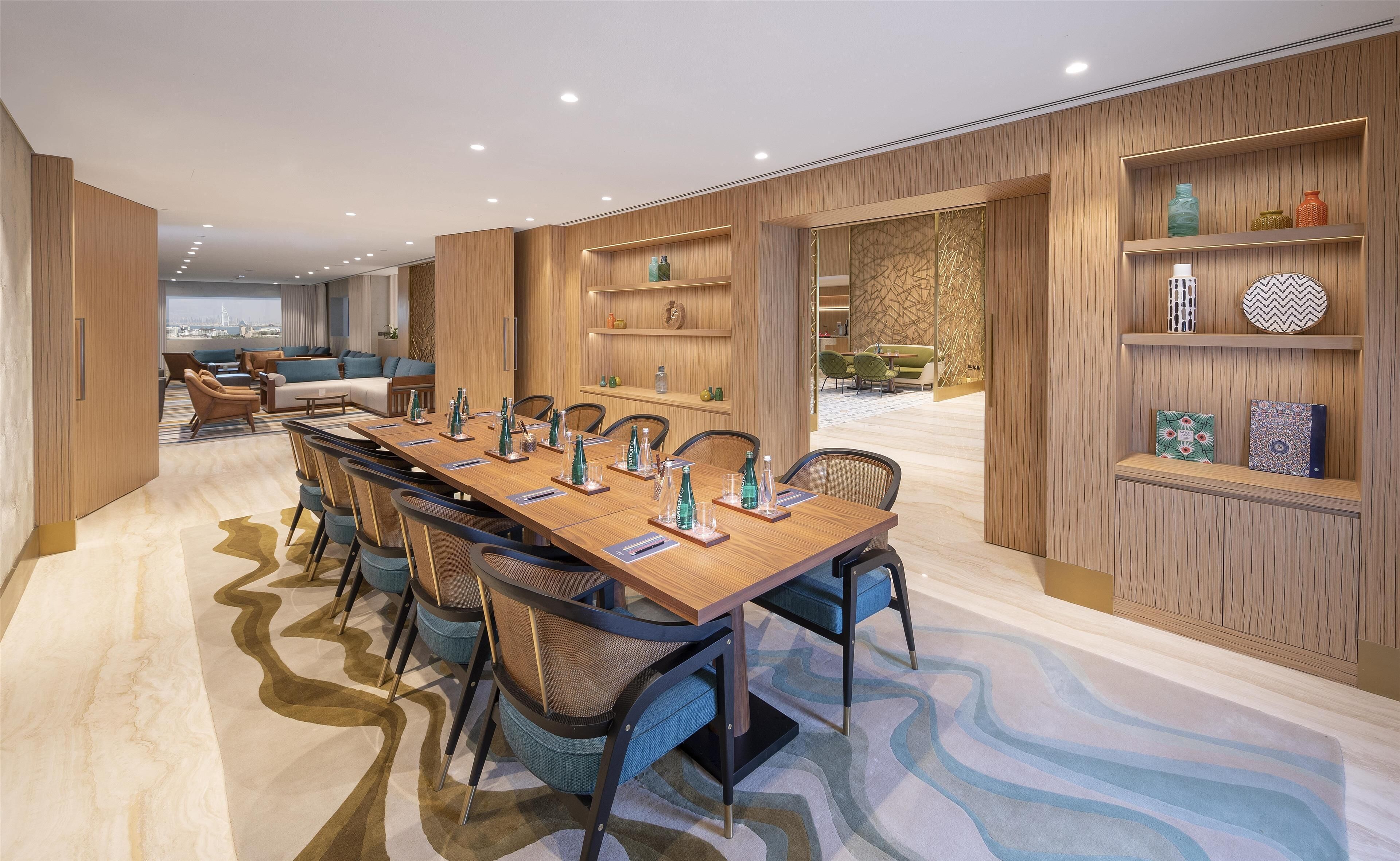 A modern meeting room with a long wooden table, blue chairs, and shelves with decorative items.