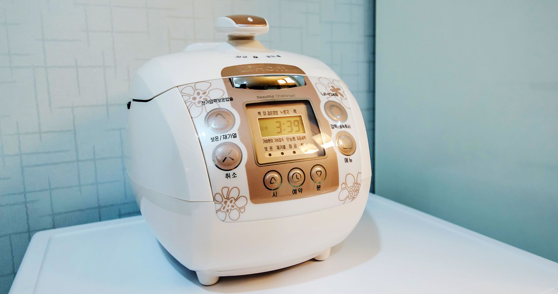 A white rice cooker with a digital display and control buttons, placed on a white surface against a tiled wall.