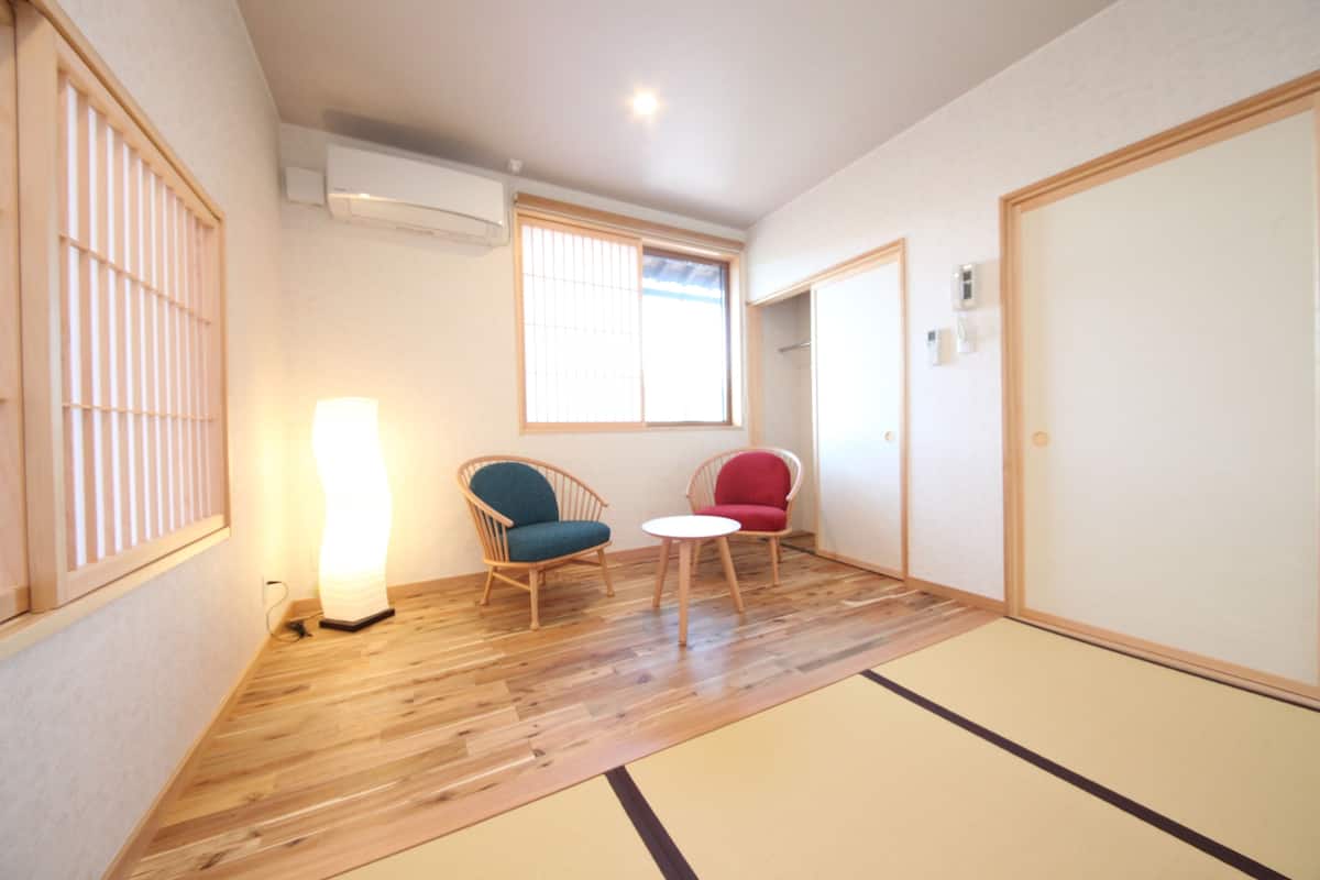 Une pièce avec un sol recouvert de tatamis, un plancher en bois, deux chaises, une petite table et une porte coulissante.