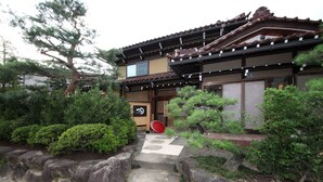 A traditional Japanese building with a tiled roof, stone steps leading to the entrance, and a well-maintained garden with various plants and a large tree. - Ichinomatsu Japanese Modern Hotel (Takayama)