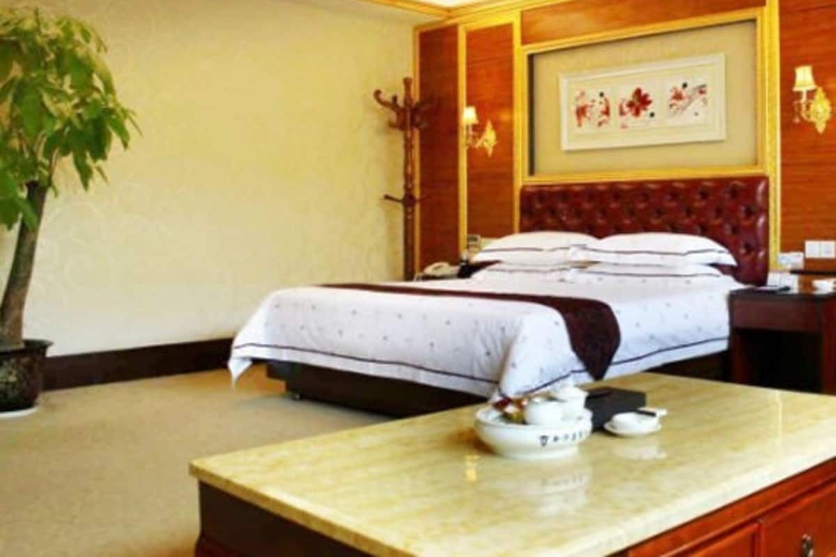 A hotel room with a bed, a nightstand, a table with a teapot, a potted plant, and a framed picture on the wall.