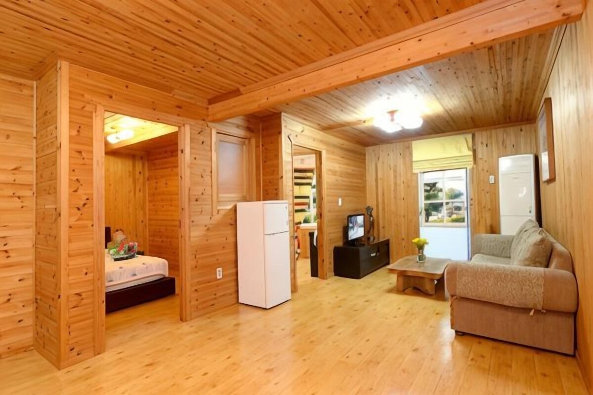 Interior de una cabaña de madera con una cama, un refrigerador blanco, un televisor y un sofá.