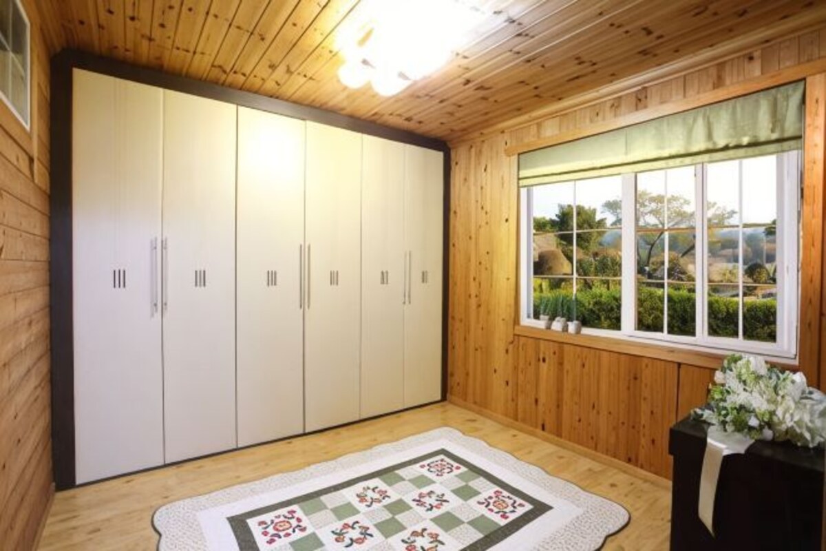 A room with wooden walls, a large wardrobe, a window with a view of greenery, and a floral arrangement on a table.