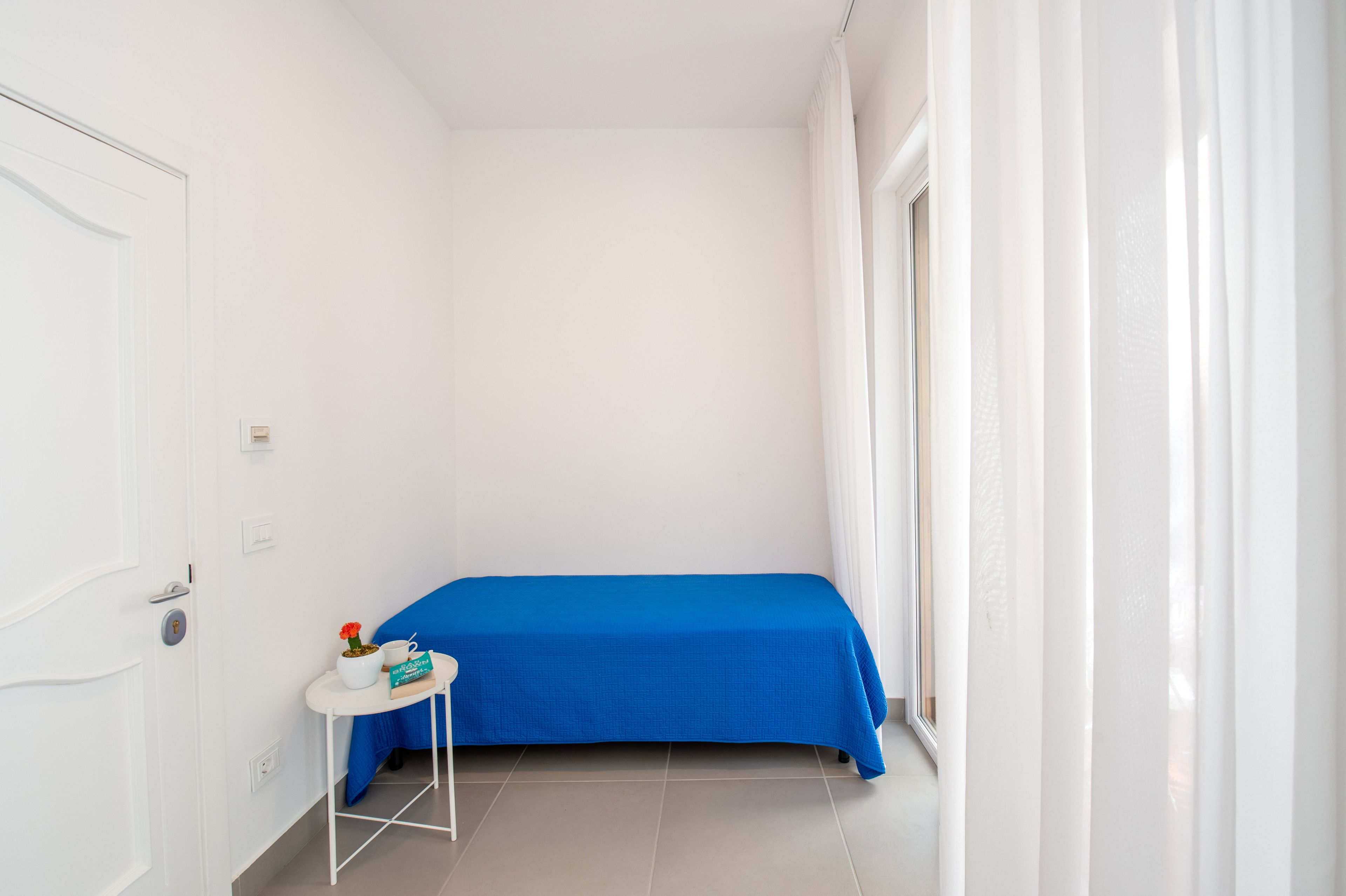 A small, bright room with a single bed, a small table with a blue tablecloth, and a window with white curtains.