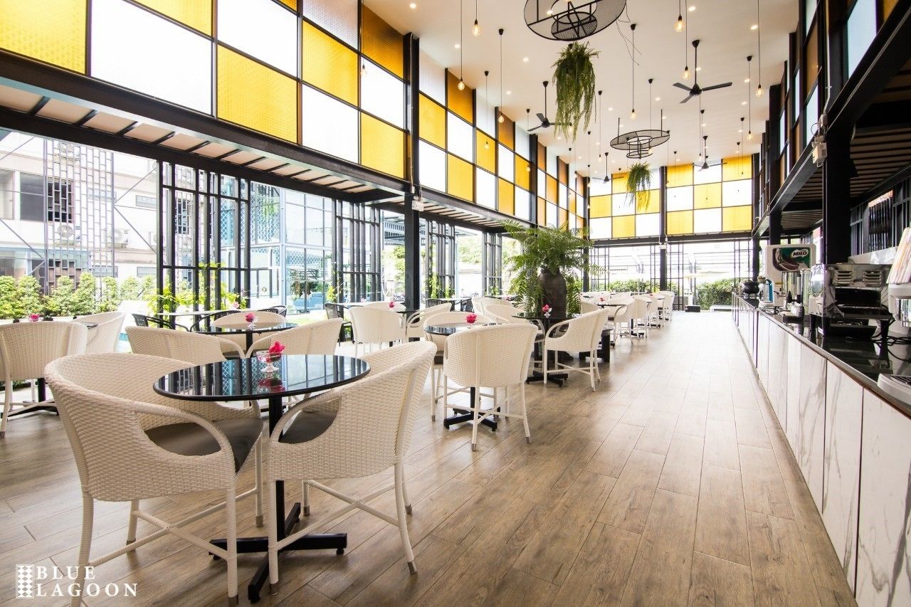 A spacious indoor dining area with round tables and white wicker chairs, large windows, and hanging plants.