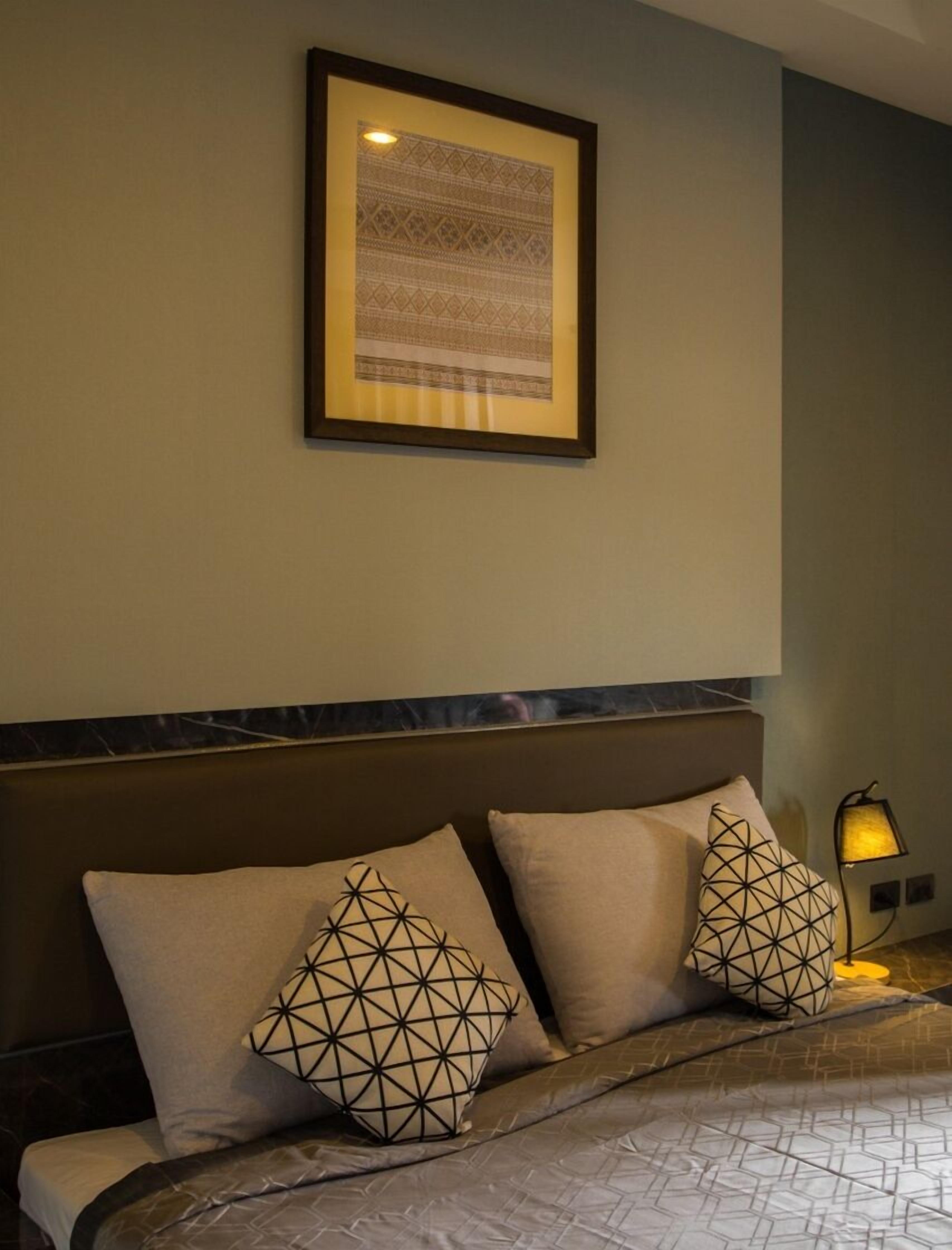A hotel room with a bed, two geometric-patterned pillows, a framed artwork on the wall, and a bedside lamp.