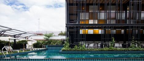 A swimming pool with a tiled deck, surrounded by greenery and a modern building with large windows.