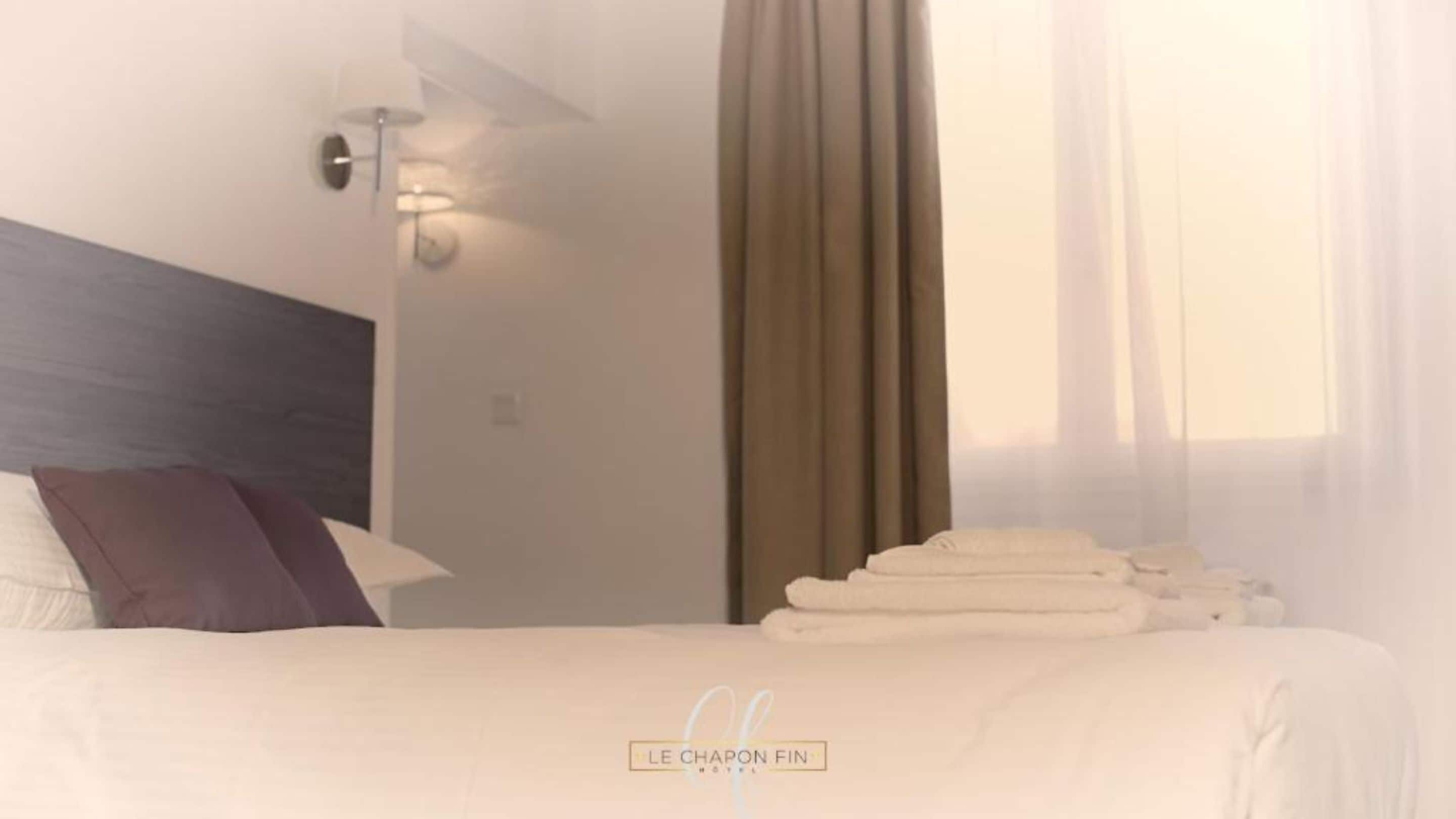 A hotel room with a bed, pillows, a lamp, a window with curtains, and a towel rack.