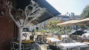An outdoor café with tables and chairs, a decorative tree, and a brick wall. - Hôtel Le Chapon Fin (Moissac)