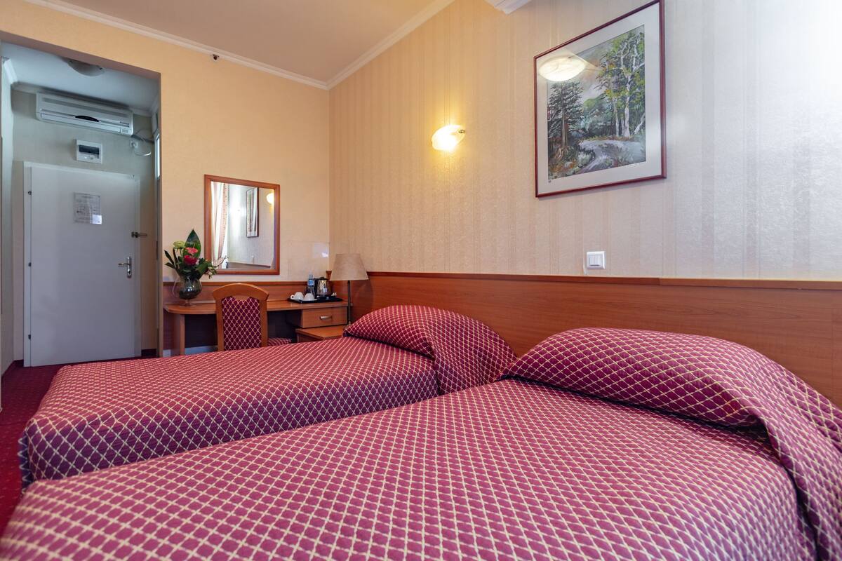 A hotel room with two beds, a desk with a chair, a mirror, and a painting on the wall.