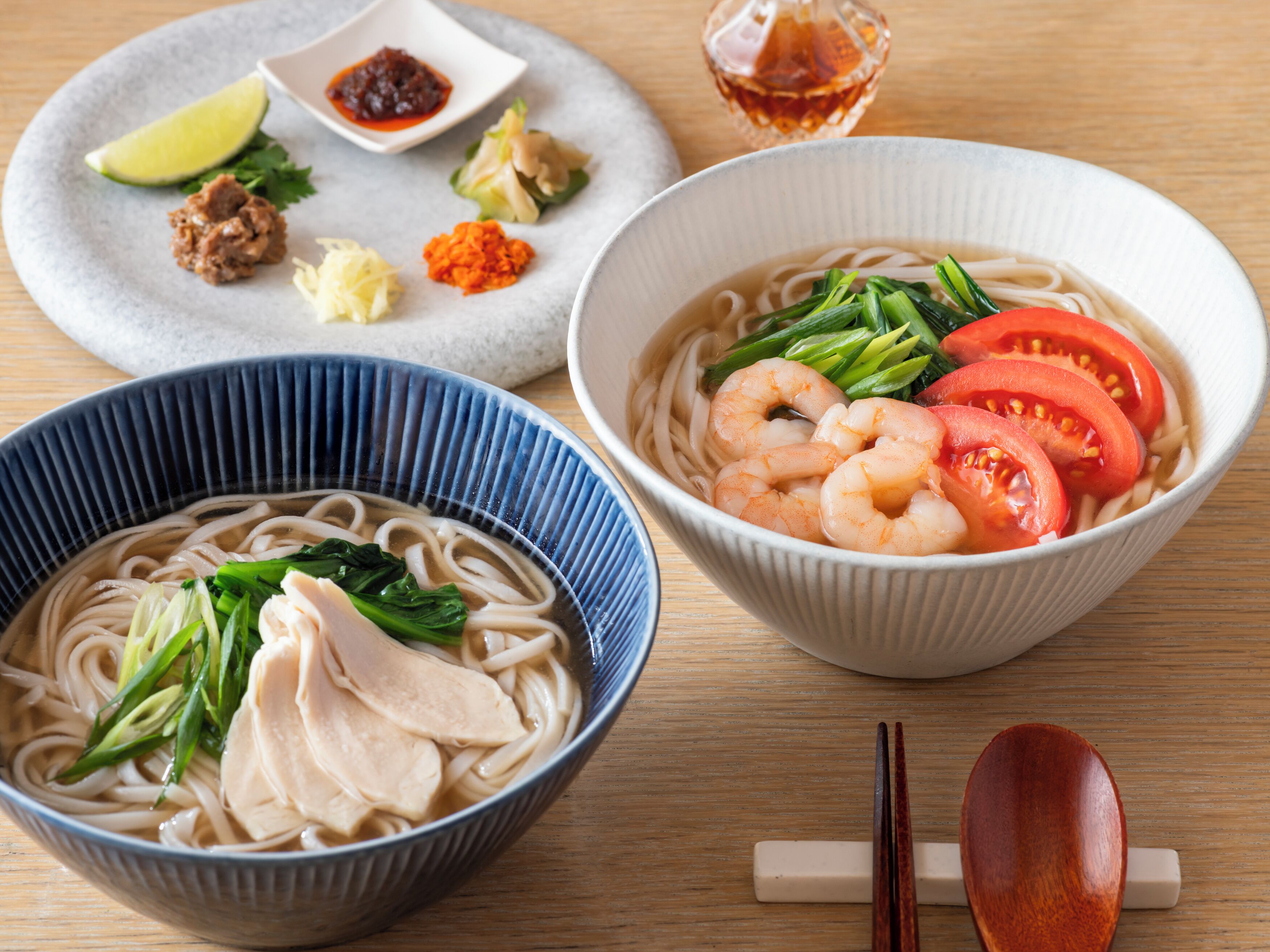 Two bowls of noodle soup with various toppings, a small dish of condiments, and a pair of chopsticks on a wooden table.