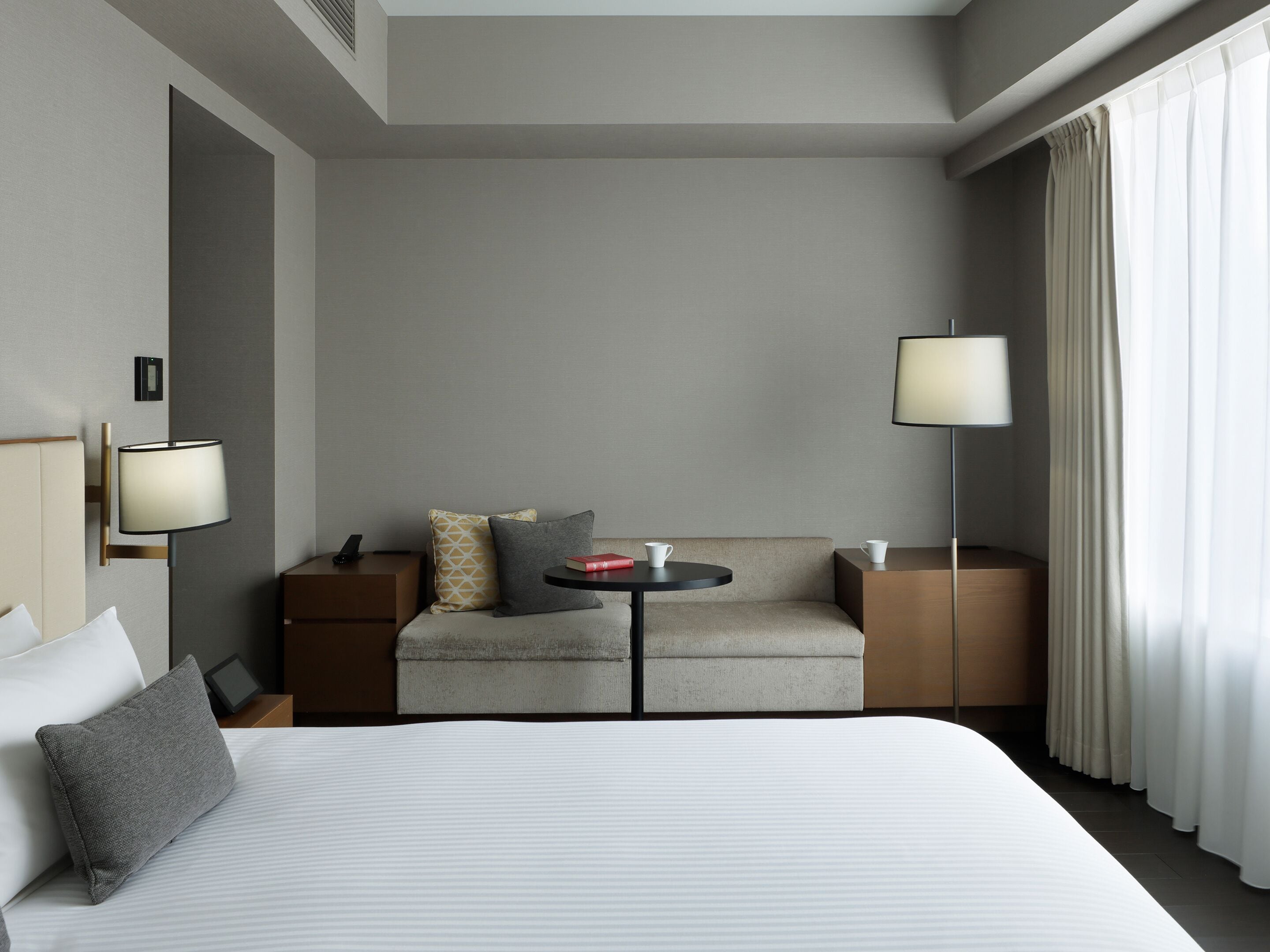 A hotel room with a bed, a sofa, a small table, two bedside lamps, and a window with curtains.