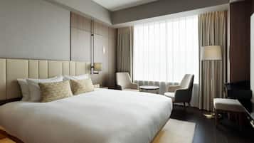 Une chambre d’hôtel avec un grand lit, deux fauteuils, une petite table ronde, un bureau avec une lampe et une grande fenêtre avec des rideaux.