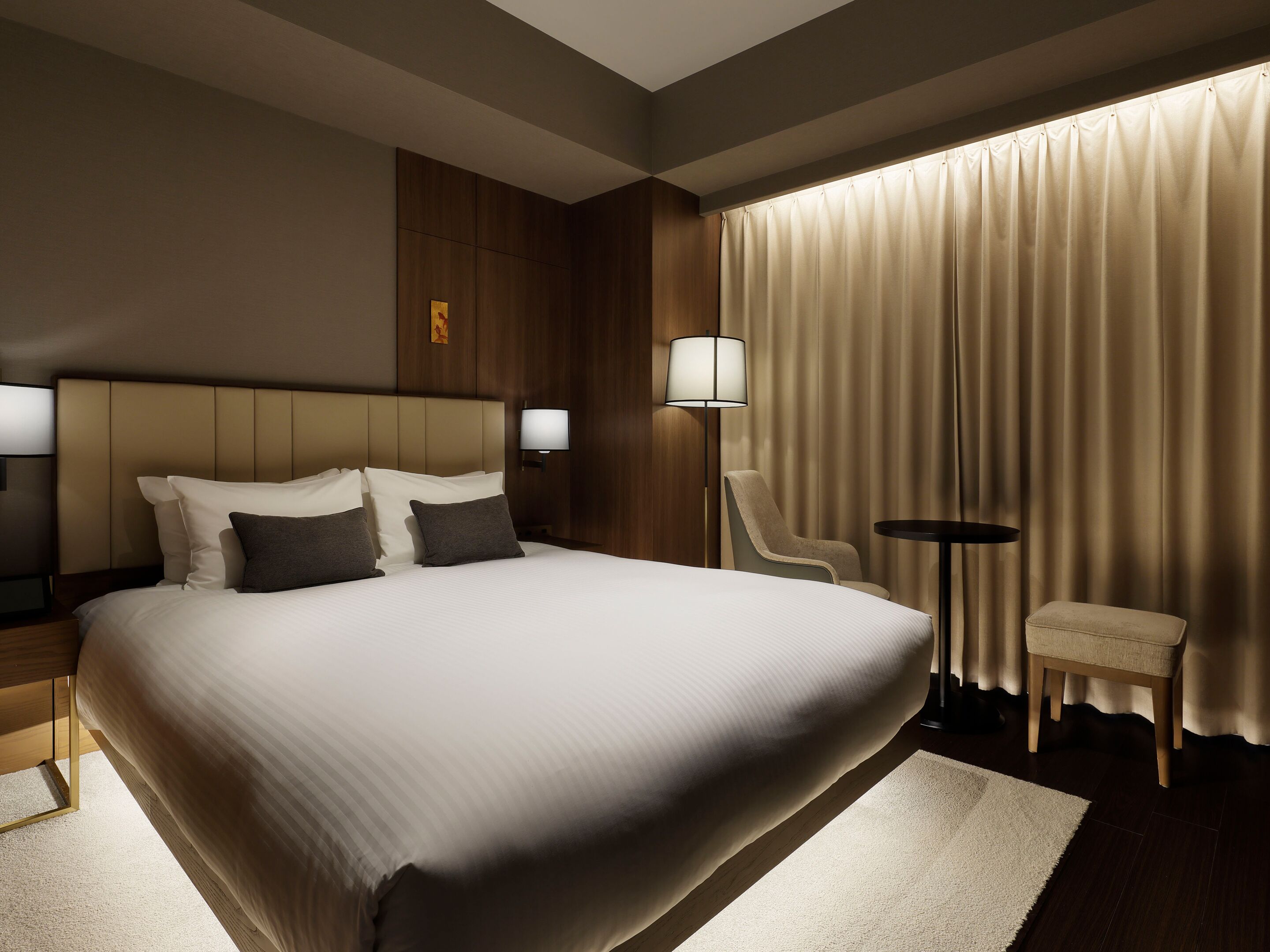 A hotel room with a large bed, two bedside tables, a chair, a small table, and a lamp.