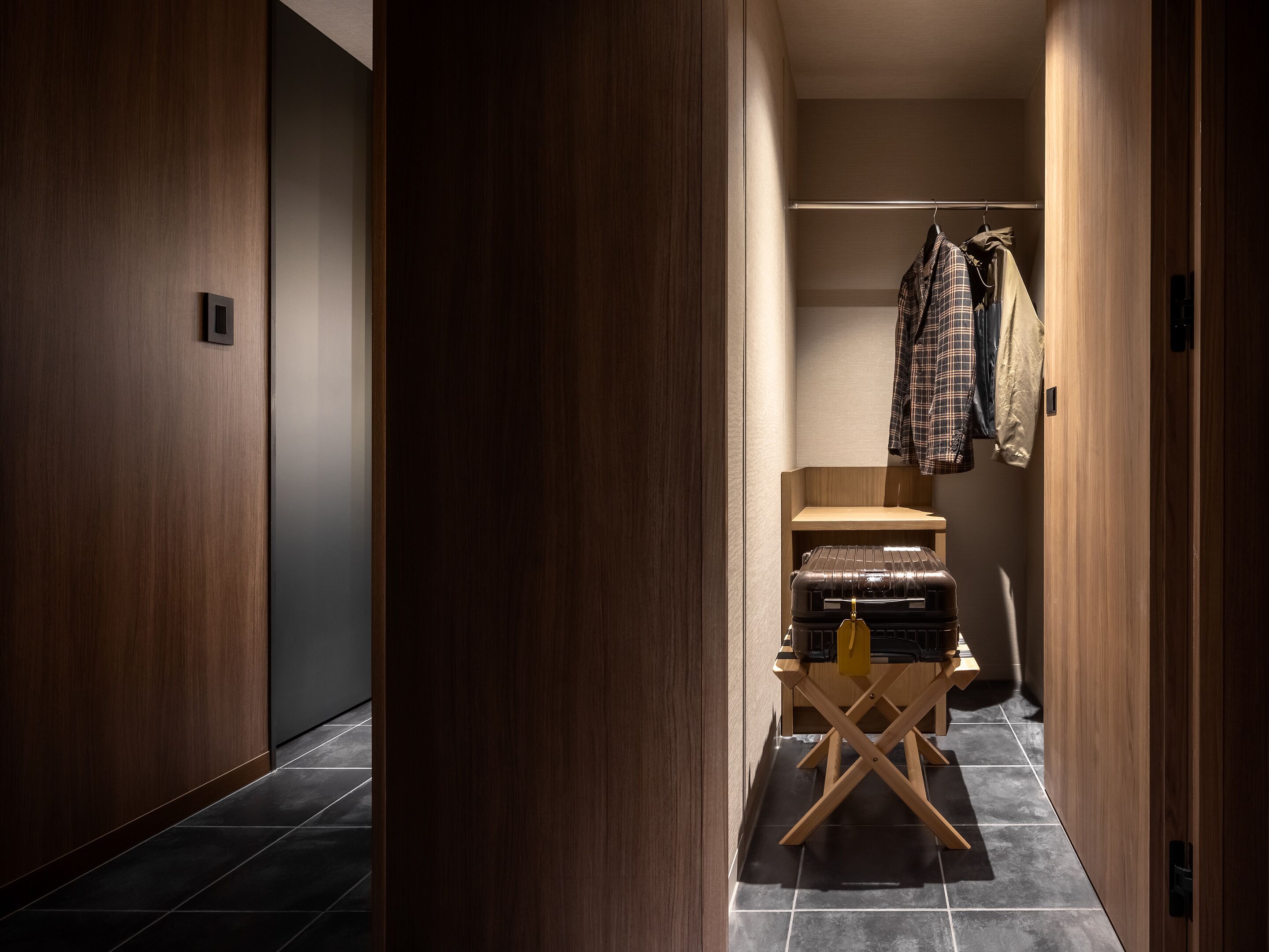 Un couloir aux murs en bois, avec un sol carrelé, un porte-manteau sur lequel sont accrochés des vêtements et une valise posée sur un présentoir en bois.