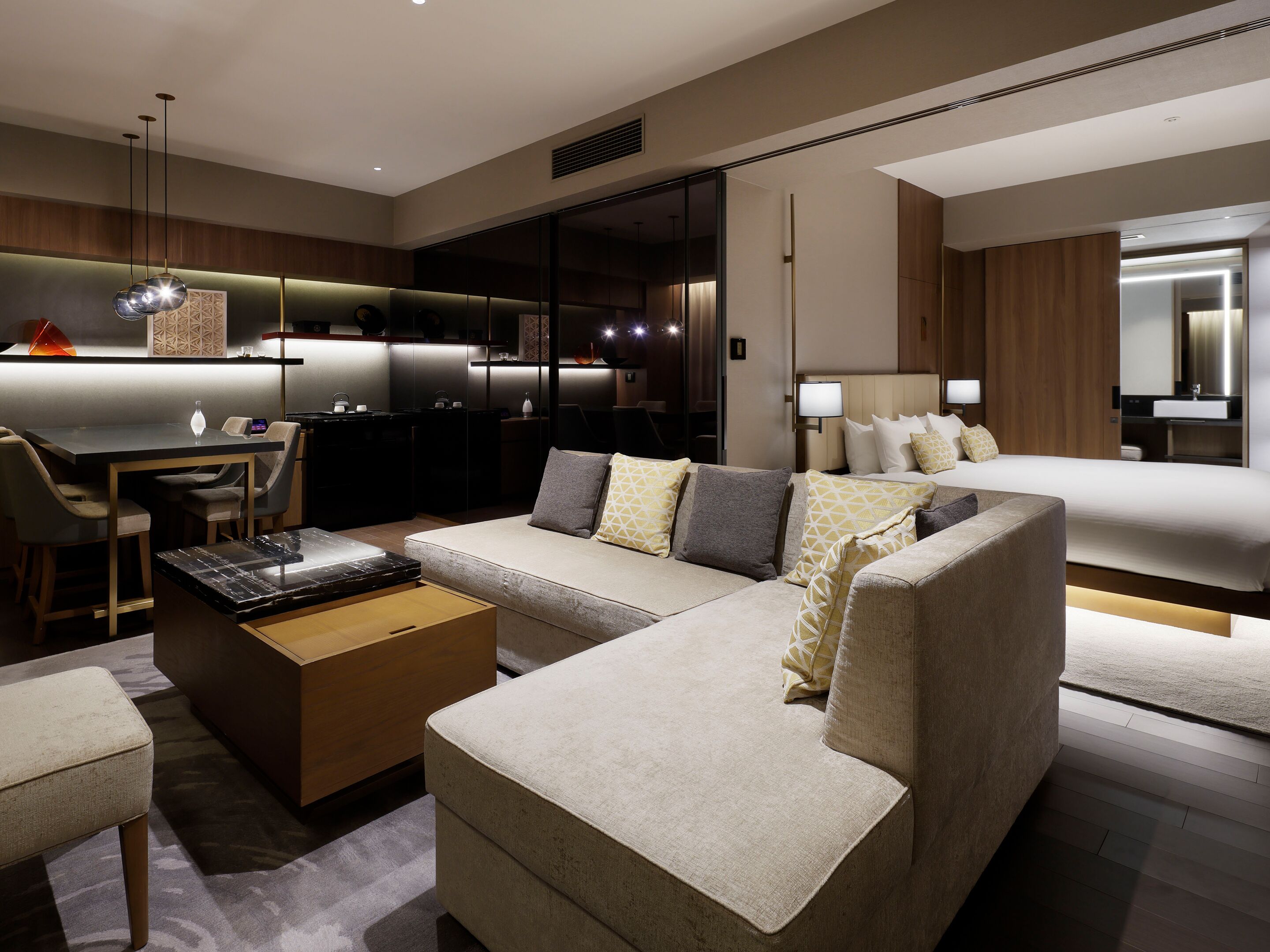 A modern hotel room with a sofa, a dining area, a kitchen, and a separate bedroom.