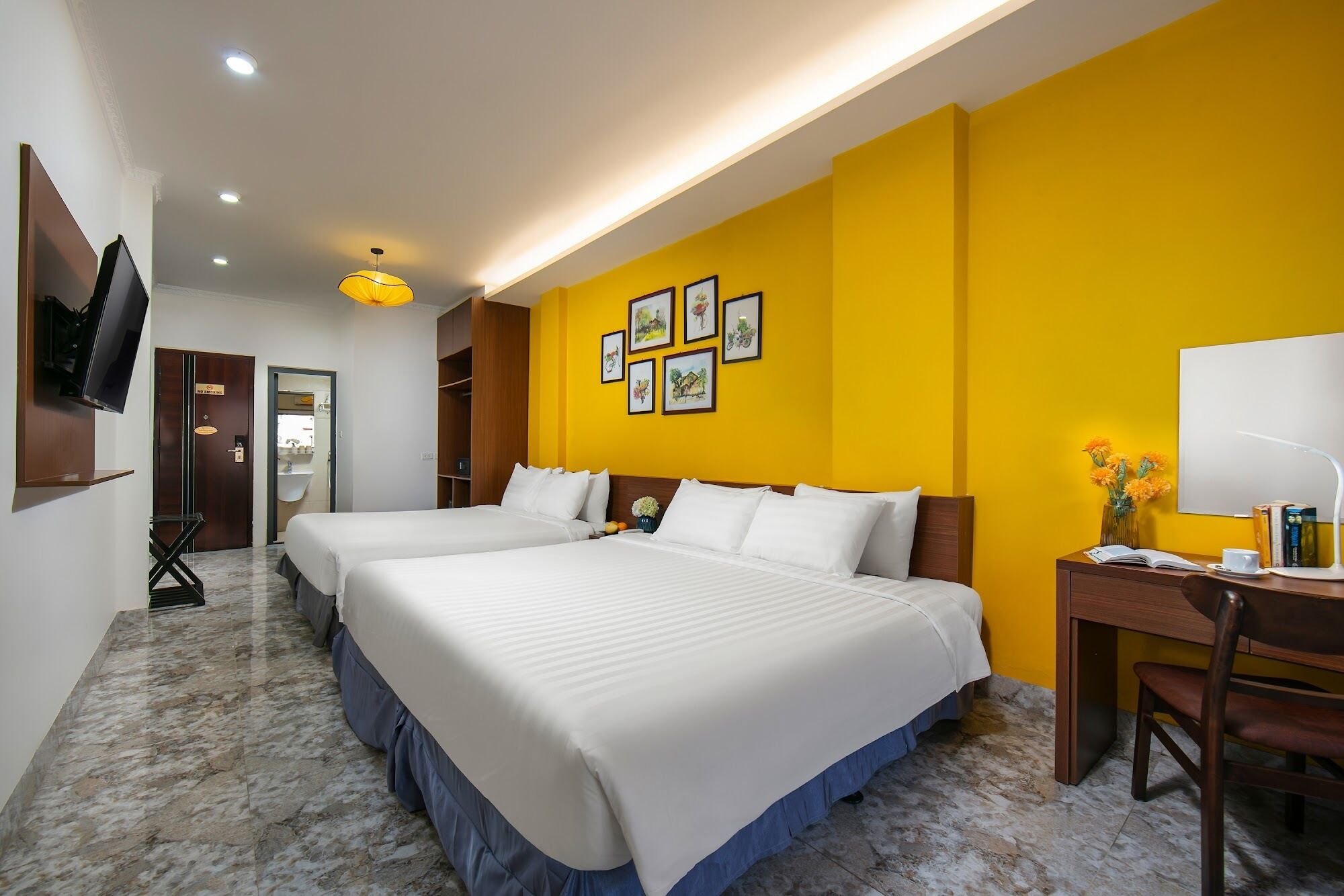 Habitación de hotel con dos camas, un escritorio, una silla, un televisor fijado a la pared y cuadros en las paredes amarillas.