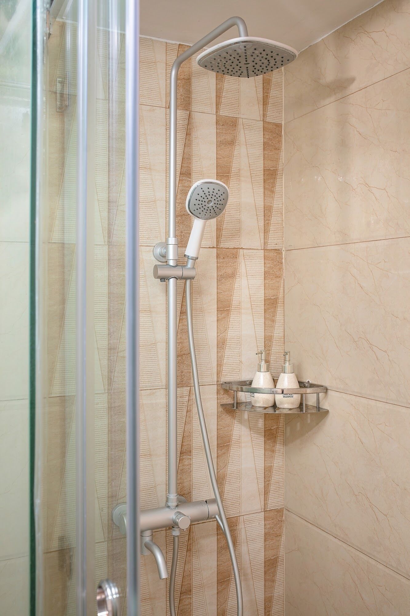 Ducha con cabezal de ducha de mano y cabezal fijo en la pared, cerramiento de ducha de cristal y estante con dos botellas de champú.