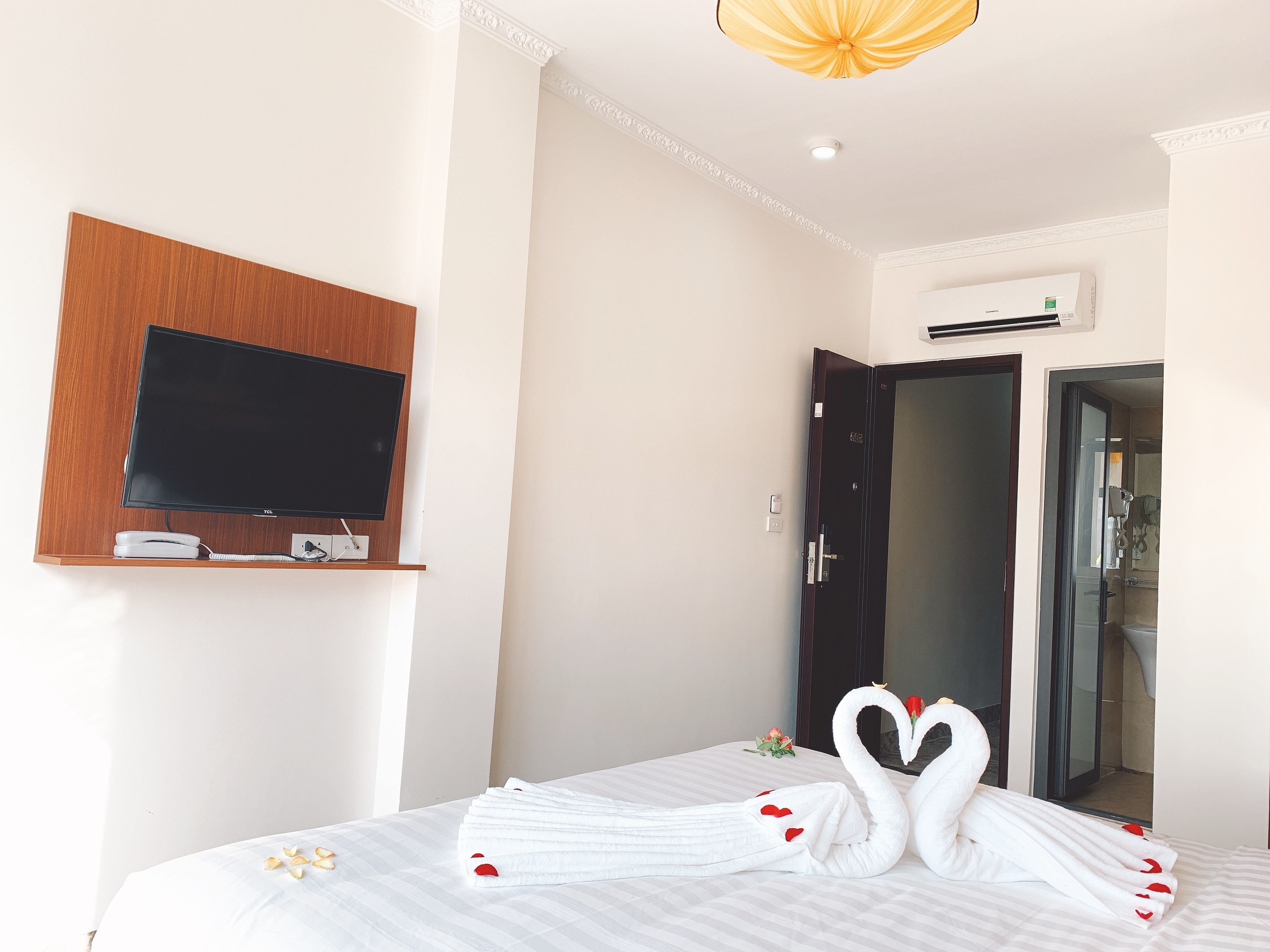A hotel room with a flat-screen TV mounted on a wooden panel, a bed with white bedding and red heart decorations, and a door leading to a bathroom.