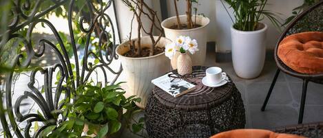 A balcony with potted plants, a wicker table with a book and glasses, a white cup, and a cushioned chair.
