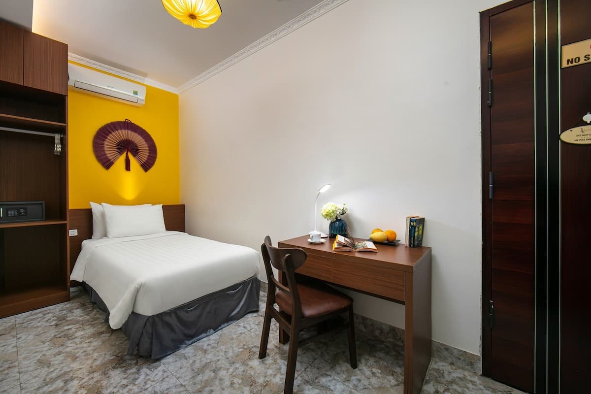 Ein Hotelzimmer mit Bett, Schreibtisch, Stuhl, Lampe, Ventilator und Tür.