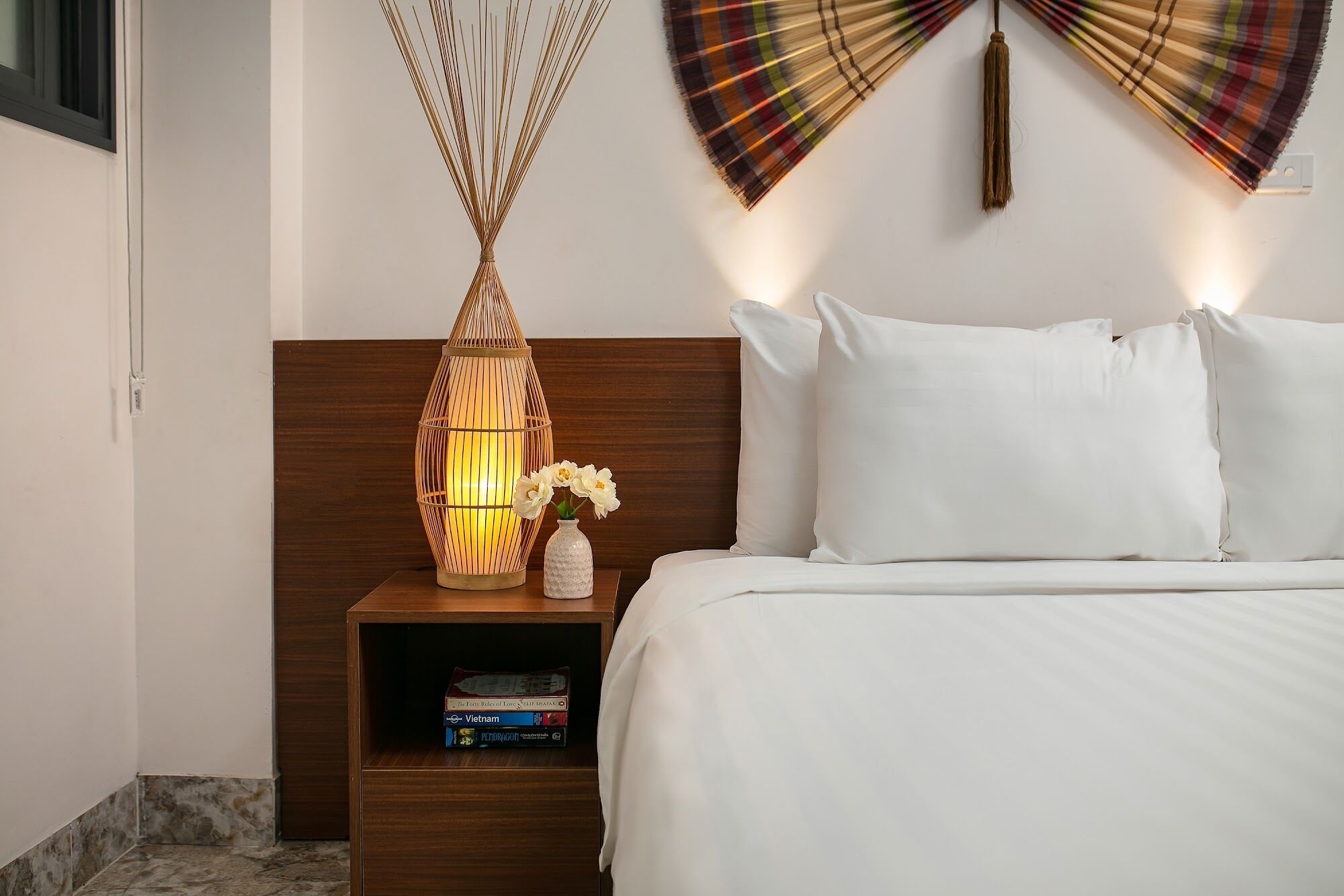 A neatly made bed with white linens, a wooden nightstand with a woven lamp, and a small shelf with books.
