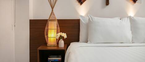 A neatly made bed with white linens, a wooden nightstand with a woven lamp, and a small shelf with books.