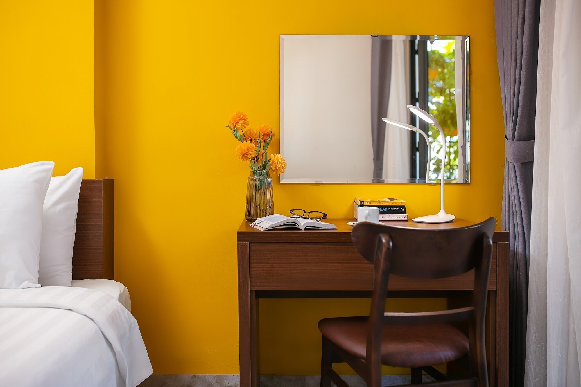 Une chambre d’hôtel avec un lit, un bureau doté d’un miroir, une chaise, un vase avec des fleurs et une lampe.