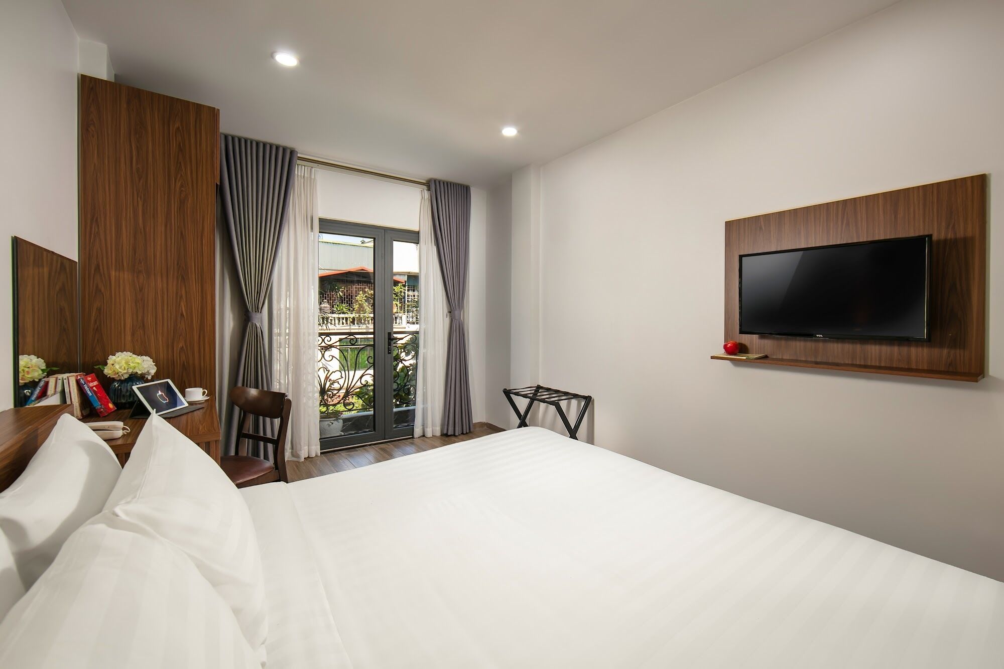 A hotel room with a bed, a TV mounted on the wall, a small table with a red apple, and a balcony with curtains.