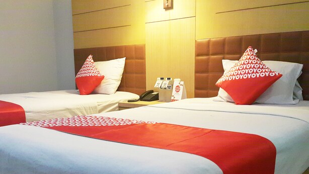 A hotel room with two beds, each with a red and white pillow, a bedside table with a phone and a tissue box, and a wall-mounted light.