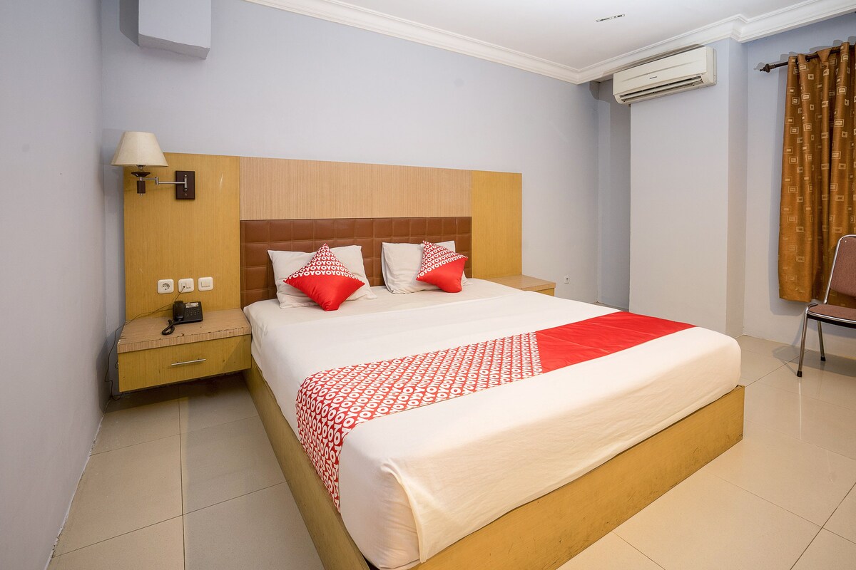 A hotel room with a large bed, a bedside table with a lamp, an air conditioning unit, and a chair.