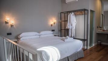 A neatly made bed with white linens, a bedside table, a wall-mounted robe rack with a white robe, and a bathroom with a sink and mirror.