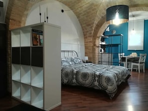 A bedroom with a patterned bedspread, a white wardrobe with shelves, a desk with a chair, and a hanging light fixture. - B&B Piccolo Loft del Borgo (Lanciano)