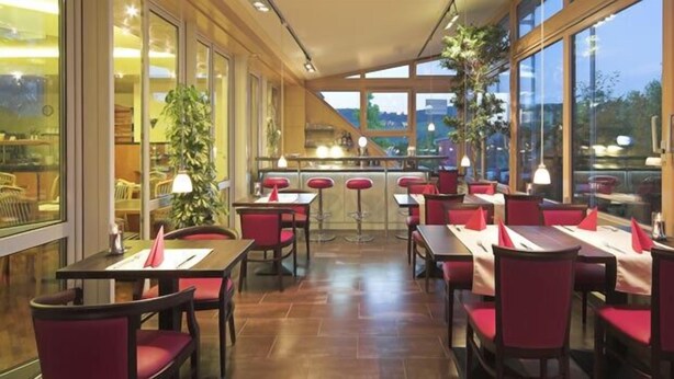 A restaurant with wooden tables and red chairs, large windows, and indoor plants. - Main Hotel Eckert (Margetshoechheim)