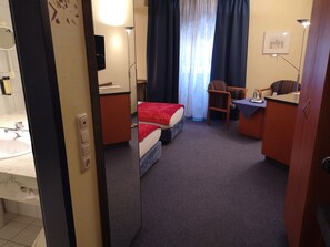 A hotel room with a bed, a desk, a chair, a small table, a lamp, a mirror, a sink, and a window with curtains. - Main Hotel Eckert (Margetshoechheim)