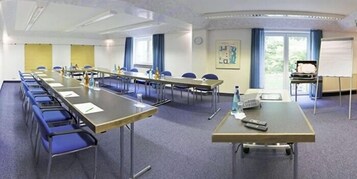 Uma sala de conferências com mesas compridas, cadeiras e uma tela de projeção.