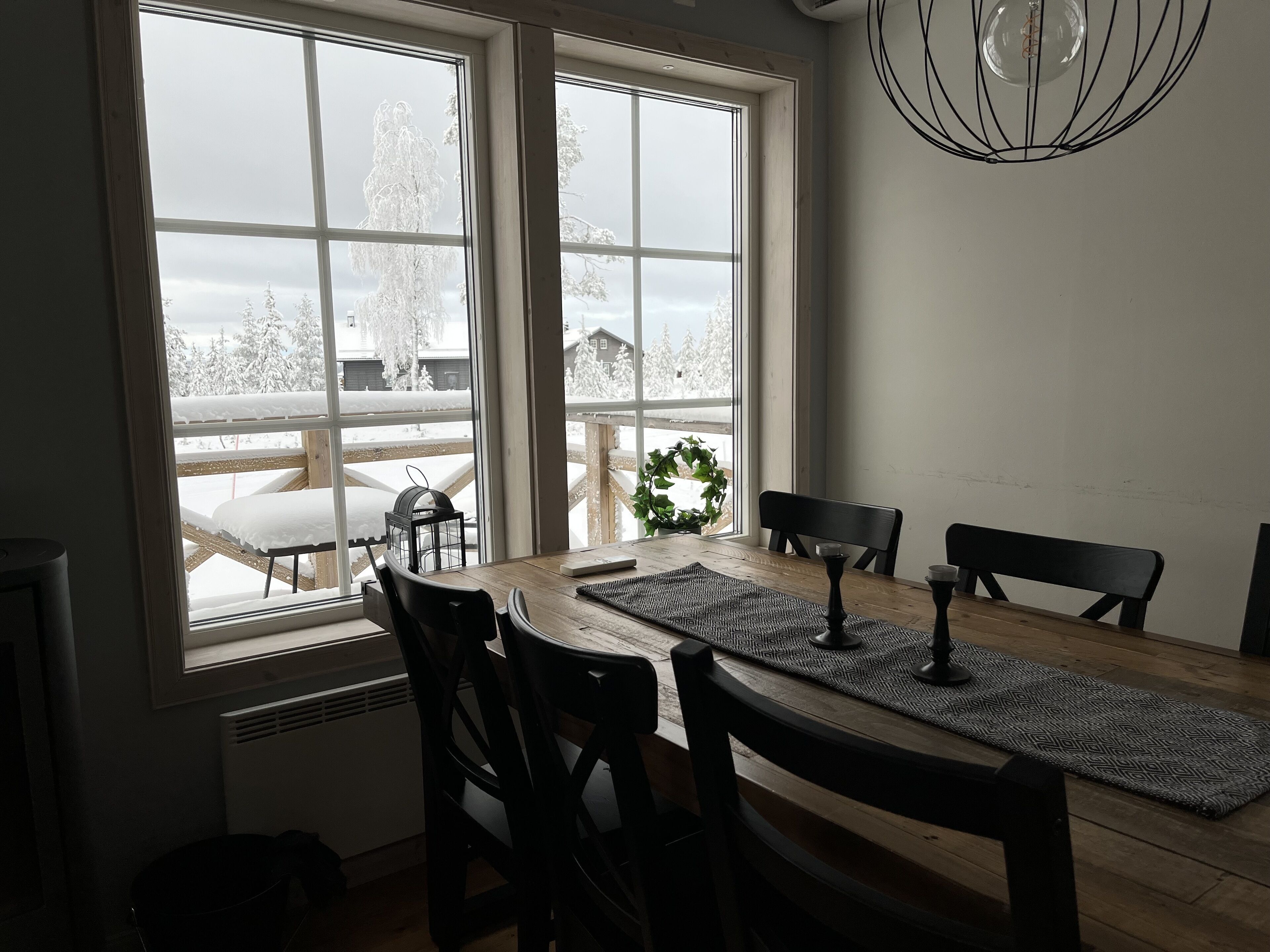 Een eethoek met een houten tafel en stoelen, grote ramen met uitzicht op een besneeuwd landschap, en een hanglamp.
