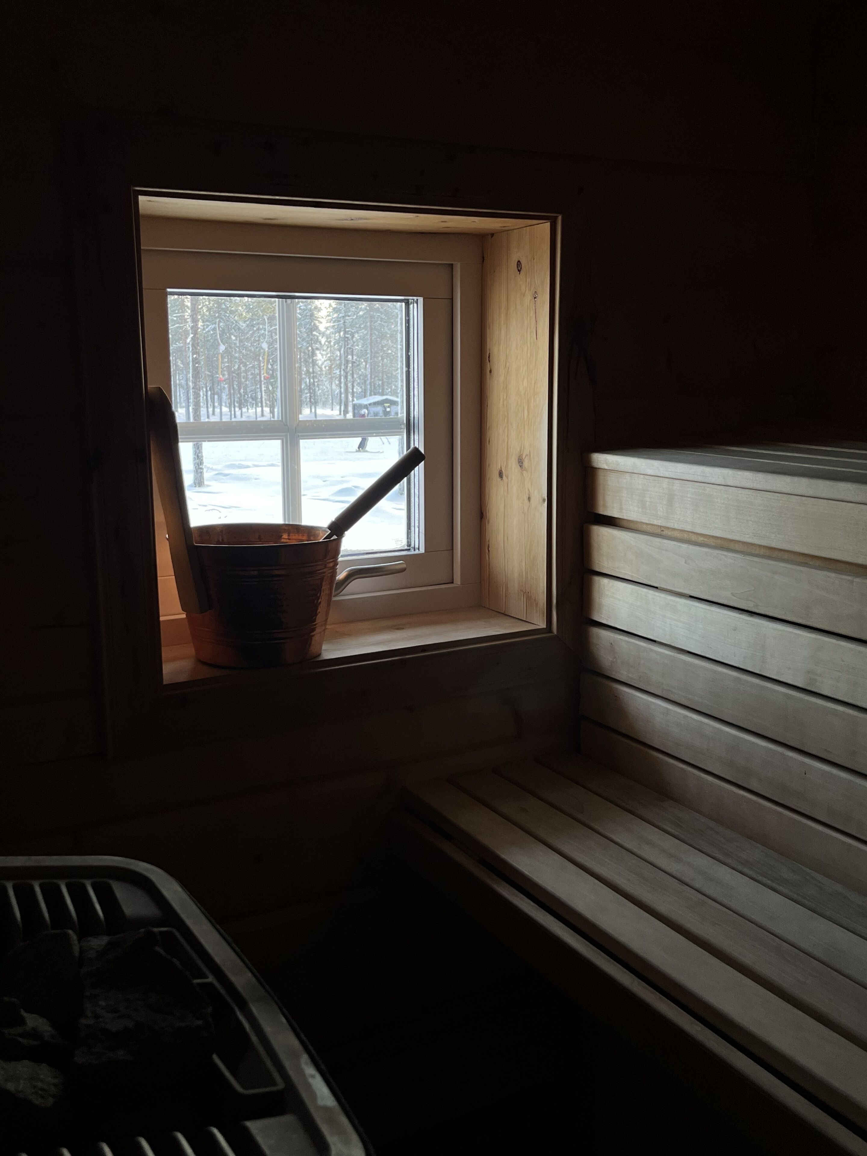 Eine kleine Holzhütte mit Fensterblick auf eine verschneite Landschaft. Im Inneren steht eine hölzerne Bank und ein Herd.