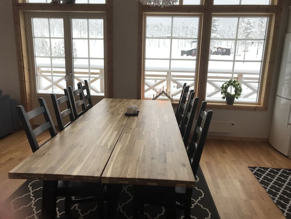 Una mesa de madera con sillas, ventanas amplias que dan a un paisaje nevado y una pequeña planta en maceta en el alféizar.