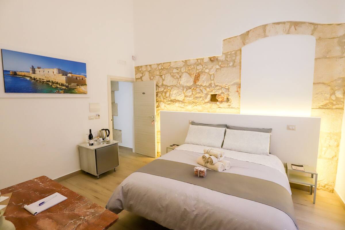 A hotel room with a stone wall, a bed with a headboard, a nightstand, a desk, and a painting of a coastal scene.