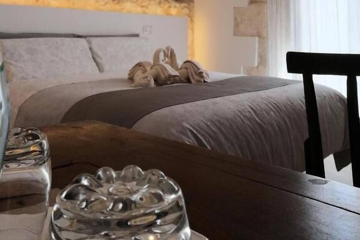 A bedroom with a stone wall, a bed with grey linens, a wooden nightstand, and a glass sculpture.